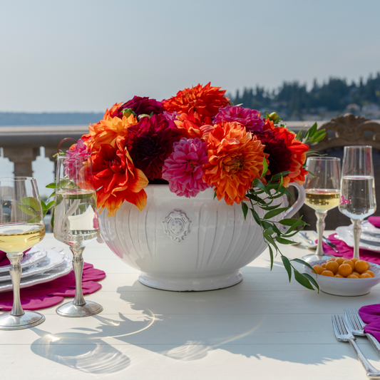 A white bella bianca garden urn filled with Maroon and orange dahlias on a white table outside on a deck