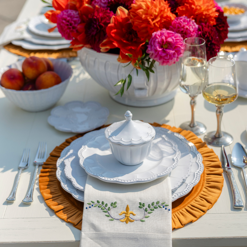Bella Bianca beaded dinnerware on a white table with Medici tri-fold napkin and orange round ruffle velvet placemat with bright flowers and wine