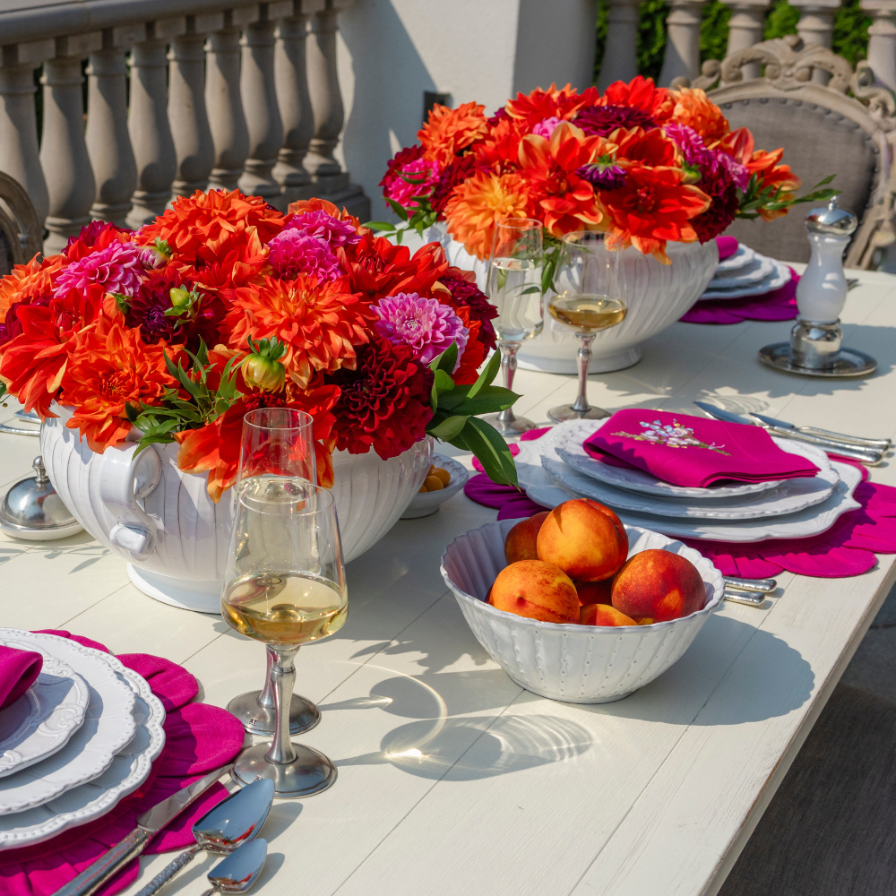 An outdoor table set with Bella Biana dishes and wine with peaches in a small serving bowl and two bright centerpieces of orange and magenta dahlias
