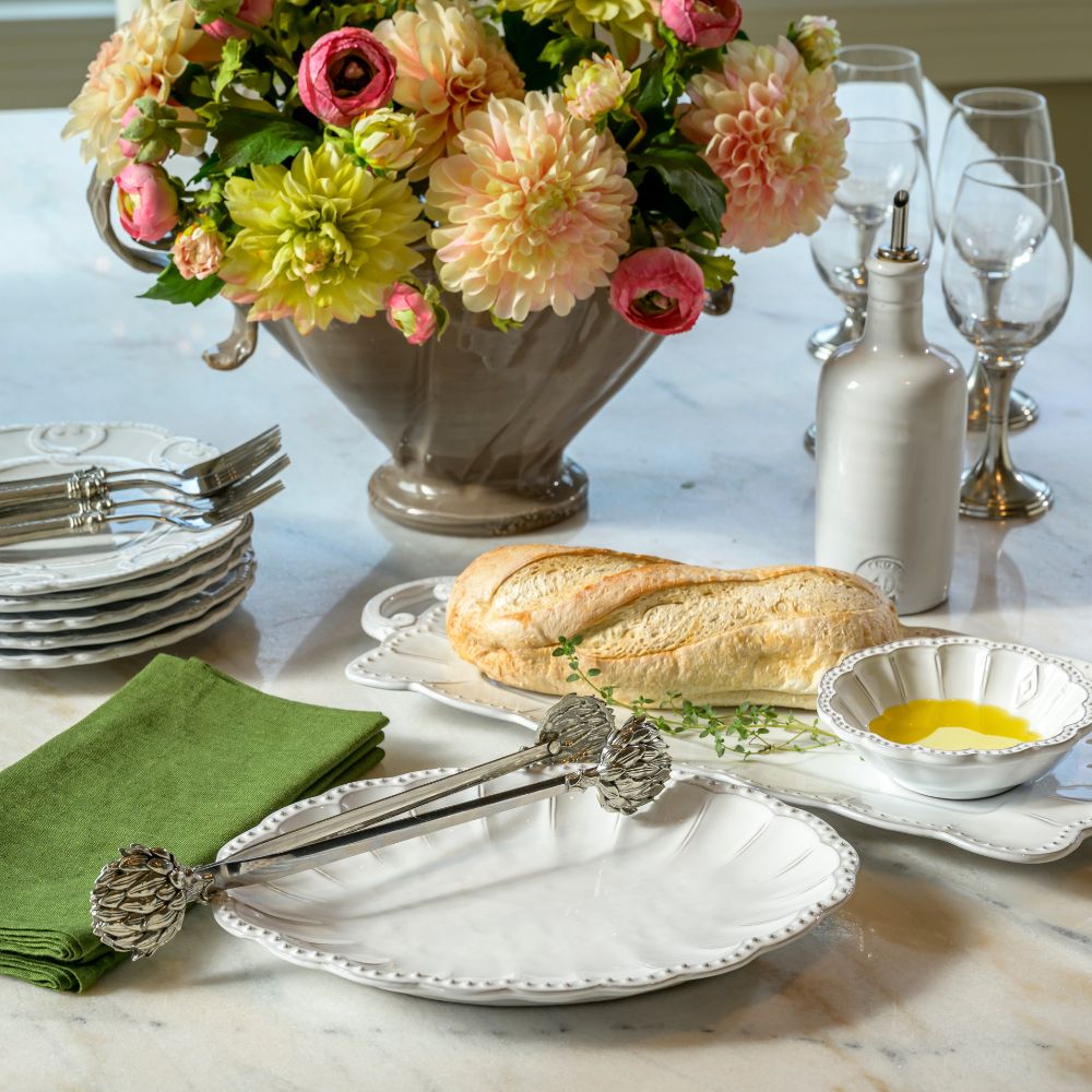 Dining table setting with bread, oil and 2 Bella Bianca platters with Artichoke Tongs with floral arrangement on a marble surface.
