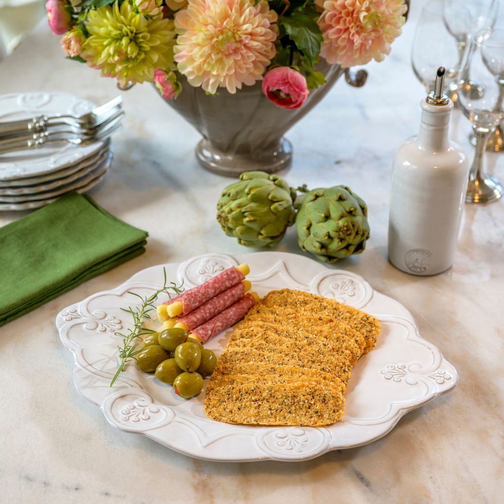 Bella Bianca Scalloped Charger used as a platter with bread, olives, and artichokes on a marble table with flowers and plates in the background.