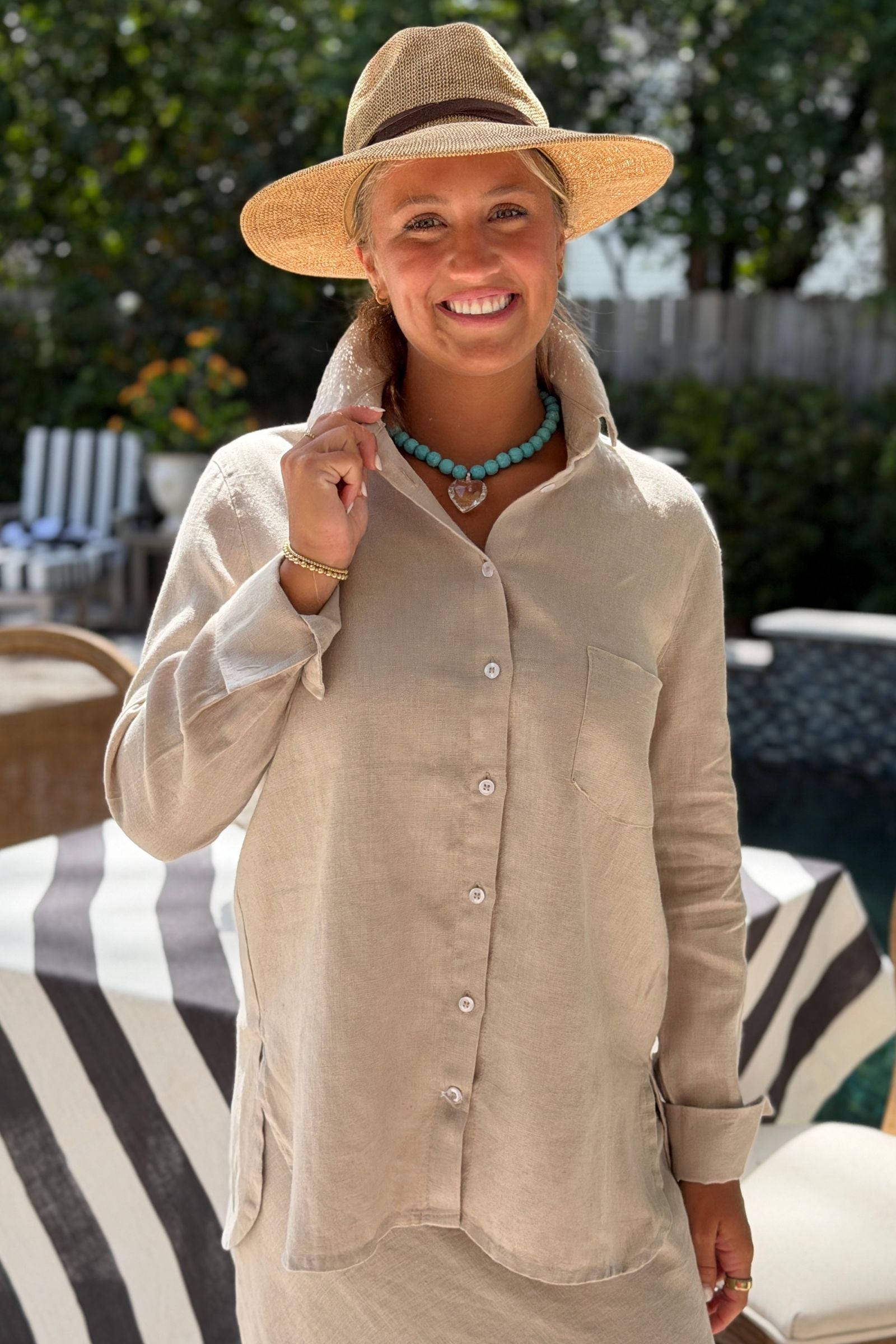 A woman wearing a soft flax linen button-up Belmont shirt, a straw hat, and a turquoise necklace, smiling outdoors in front of a patio setting.