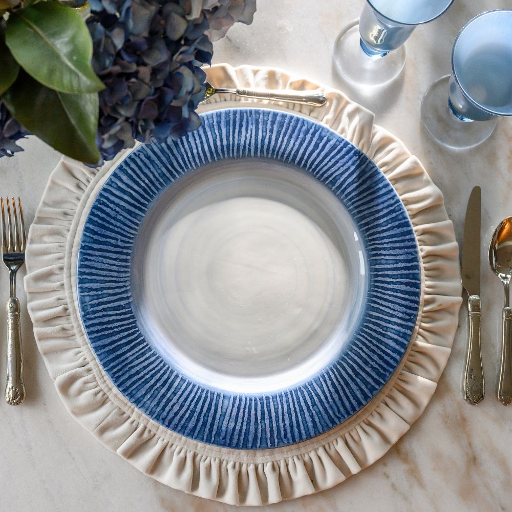 Table setting with a blue  and cream striped charger, silverware, and floral arrangement on a marble surface.