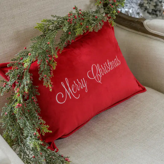 A red velvet boudoir pillow with 'Merry Christmas' written in white script, displayed on a beige chair with green foliage decoration.