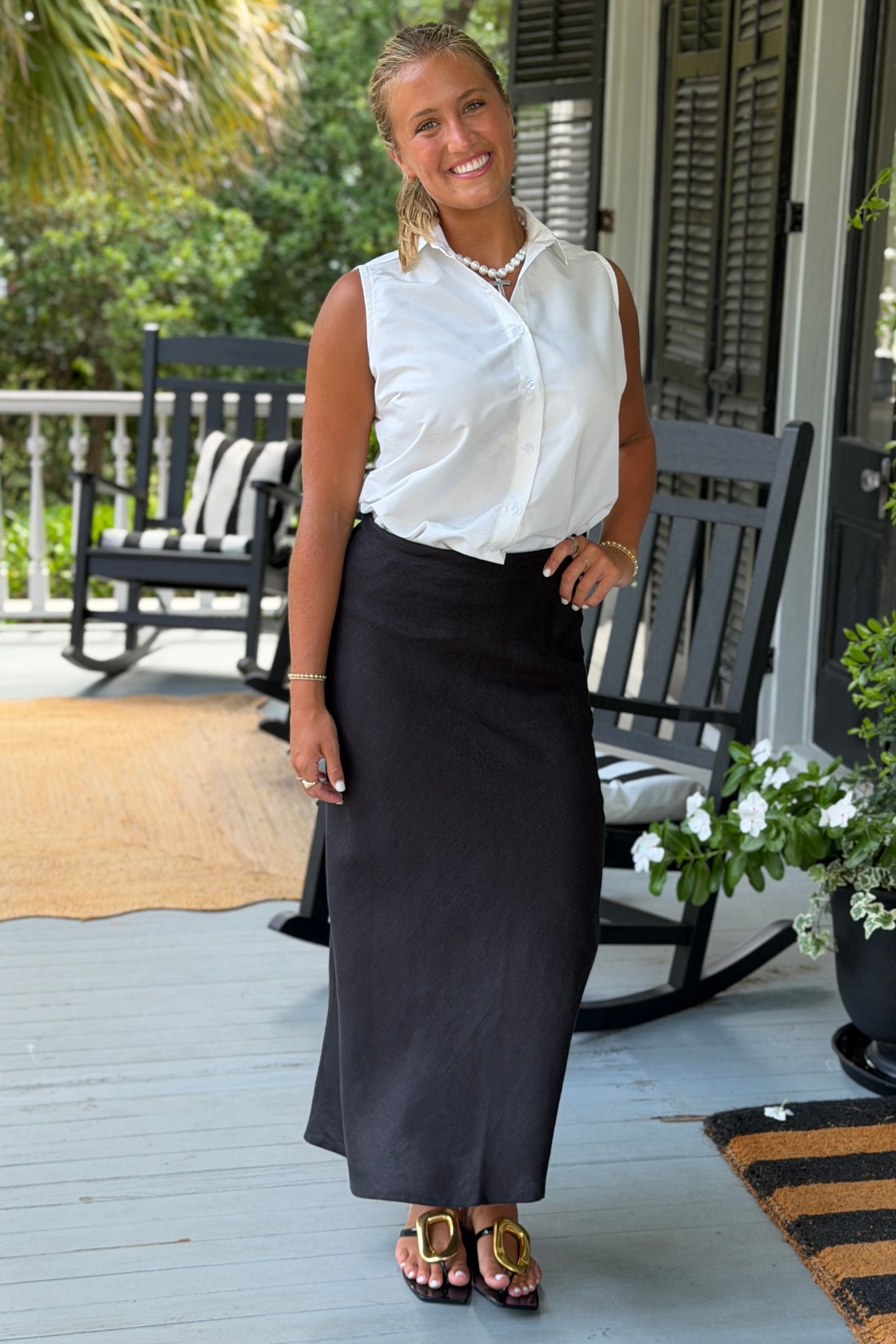 A woman stands on a porch wearing a sleeveless white button-up Caroline Taffeta blouse and a maxi Cindy black linen skirt, with gold sandals and a pearl necklace. The background shows a home porch with rocking chairs, greenery, and shutters.