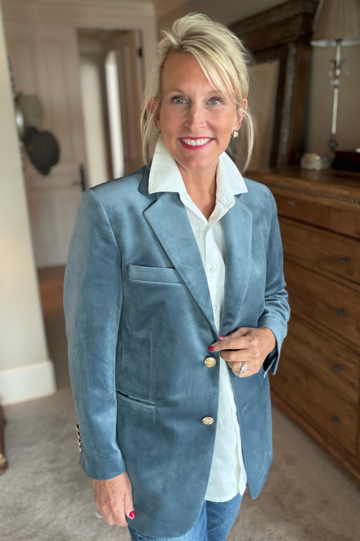 Charlie Velvet Blazer in French blue with gold button detail, styled over a white collared shirt with jeans in an elegant bedroom setting