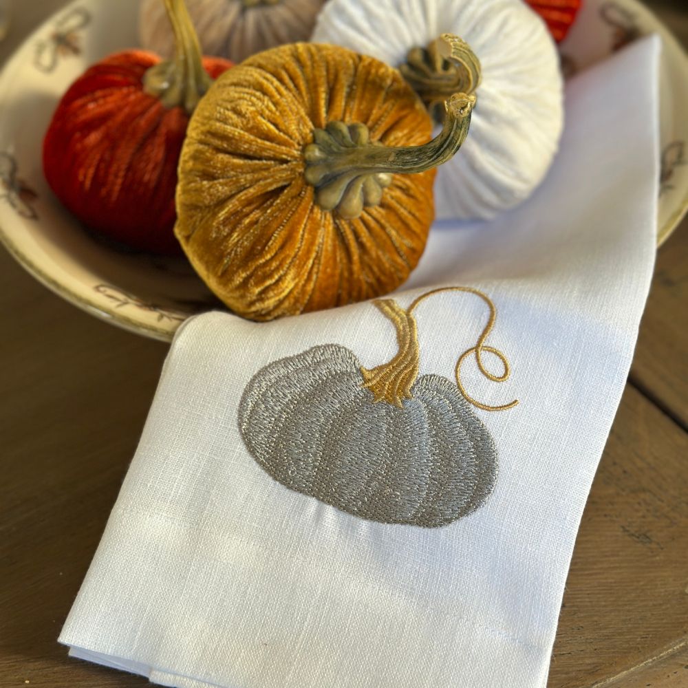 Embroidered cinderella pumpkin design on a white linen tea towel with decorative pumpkins in the background.
