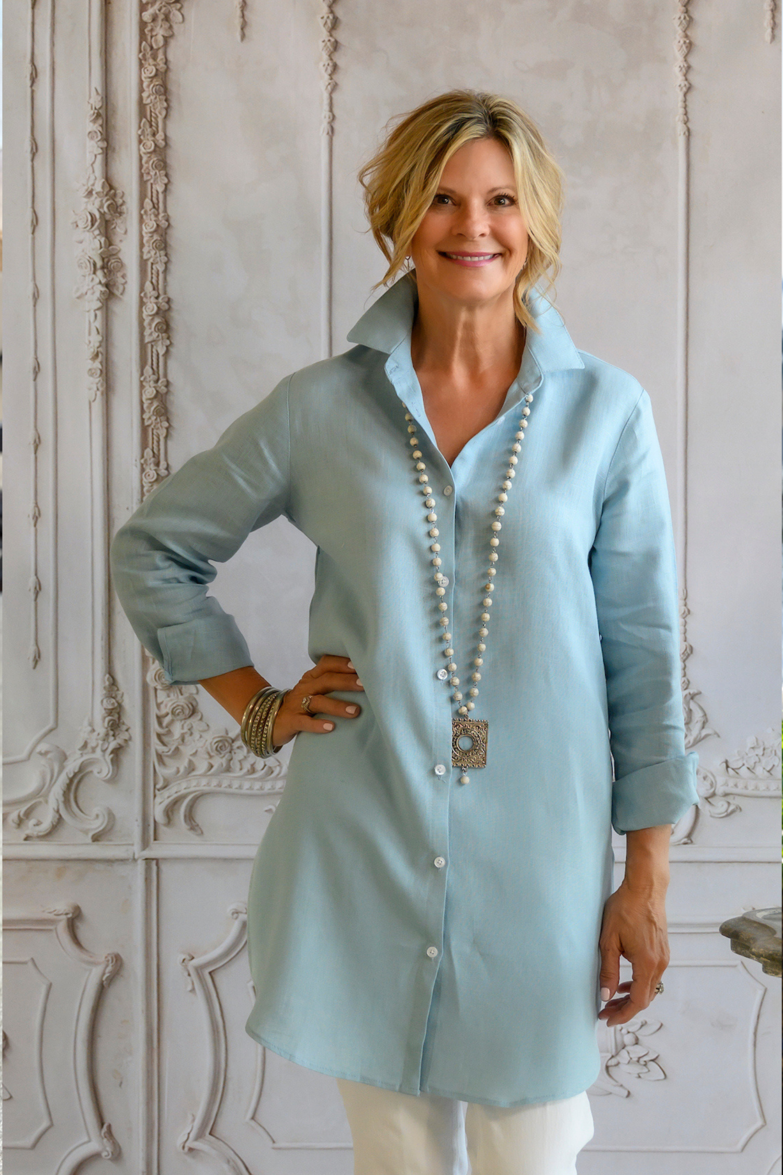 Woman wearing light blue linen Christina button up tunic aired with white jeans, boho beaded necklace, and pewter bangles. --Arte Italica and Crown Linen Designs