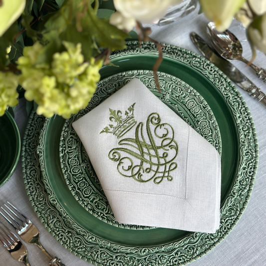 A white linen napkin embroidered with a green crest design folded on a renaissance ceramic plate on a table with flowers