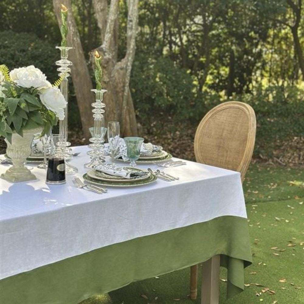 Elegant outdoor table setting with white linen tablecloth and green accents, surrounded by trees.