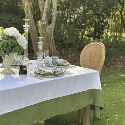Elegant outdoor table setting with white linen tablecloth and green accents, surrounded by trees.