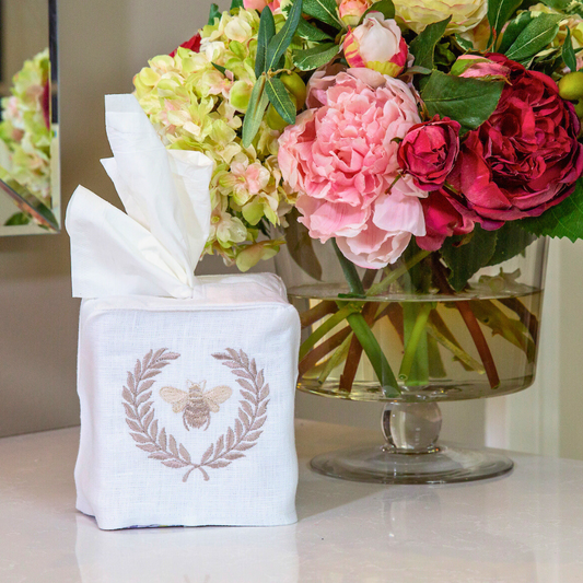 A white linen tissue box cover with a Taupe embroidered bumble bee design, displayed in front of a floral arrangement.