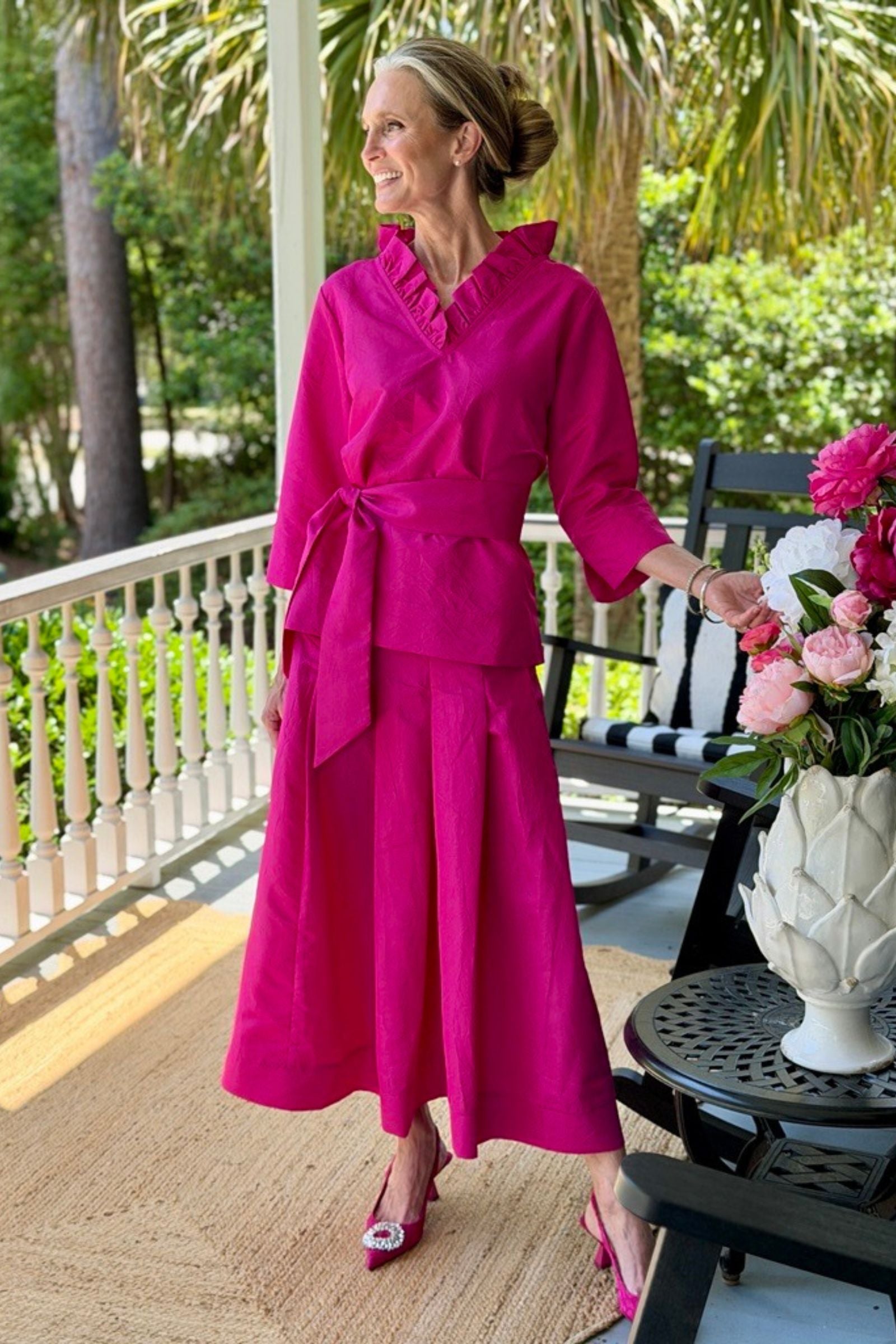 Woman in a bright fuchsia Darcy top and Lizzy Skirt standing on a porch with greenery in the background