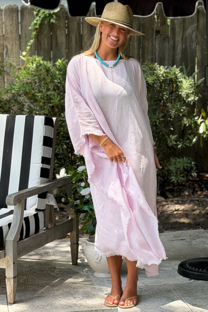 A woman wearing a Lilac lightweight Eden kaftan dress, a straw hat, and a turquoise necklace stands on a patio in front of a wooden fence and greenery.