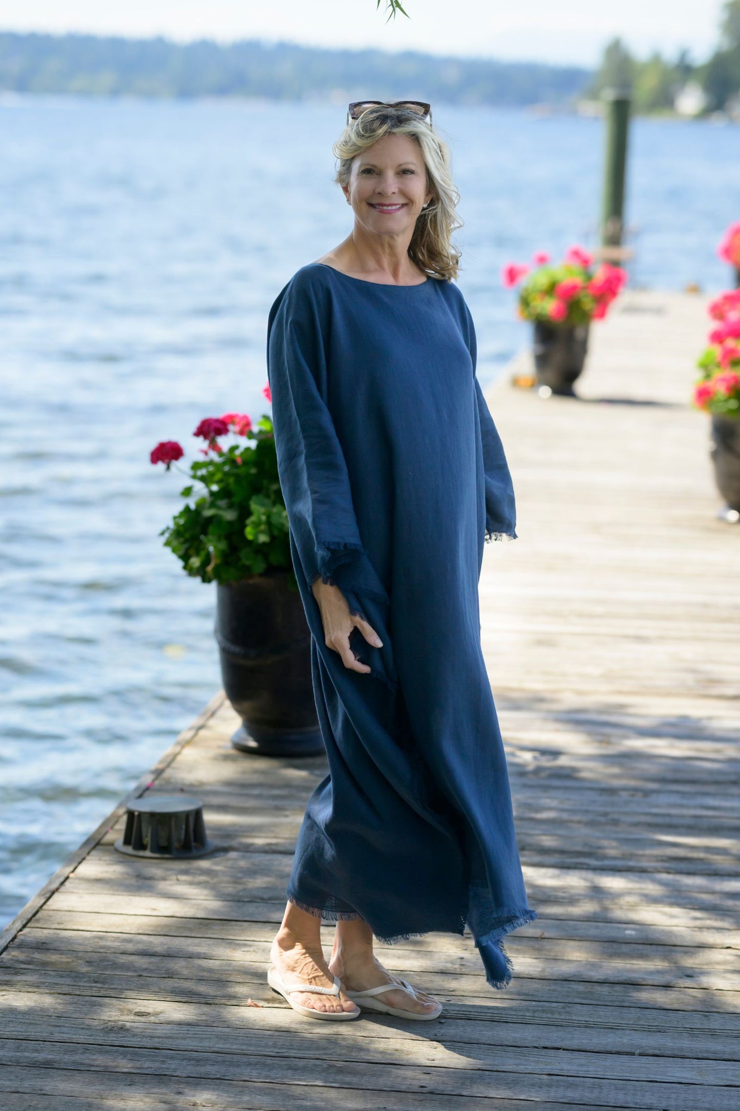 Woman in a navy blue linen kaftan  standing on a wooden dock by a lake with flowers and potted plants.