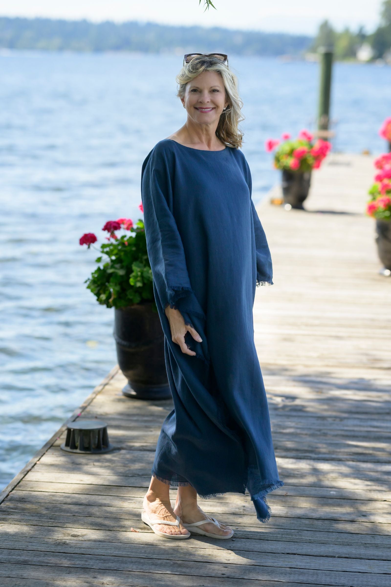 Woman in a navy blue linen kaftan  standing on a wooden dock by a lake with flowers and potted plants.