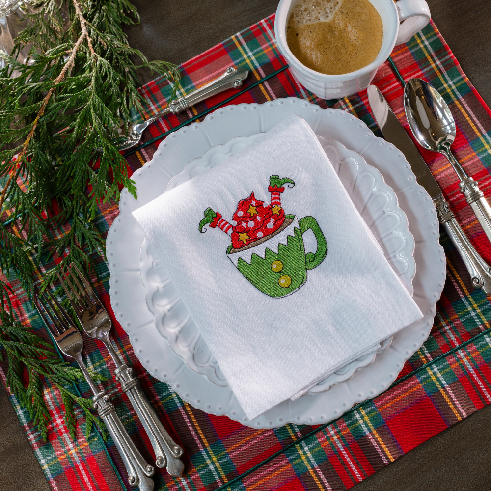 A Bella Bianca placesetting on a red tartan cotton placemat with an embroidered elf in a cup white linen towel folded on it