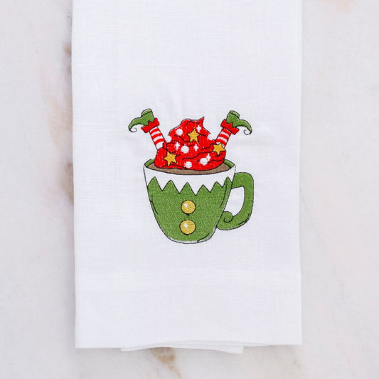 White linen towel with an embroidered design of an elf with Red socks sticking out of a green mug on a white background