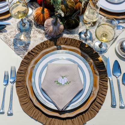 An large flax linen napkin embroidered with a white pumpkin and greenery folded on a Fall table with Tuscan dinner plates and a brown velvet round ruffle placemat