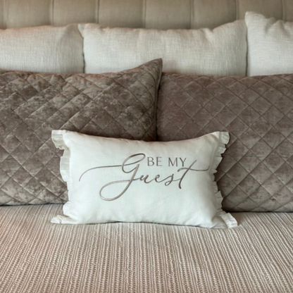 Decorative pillow in 100% linen with 'Be My Guest' embroidered text on a bed with small ruffles