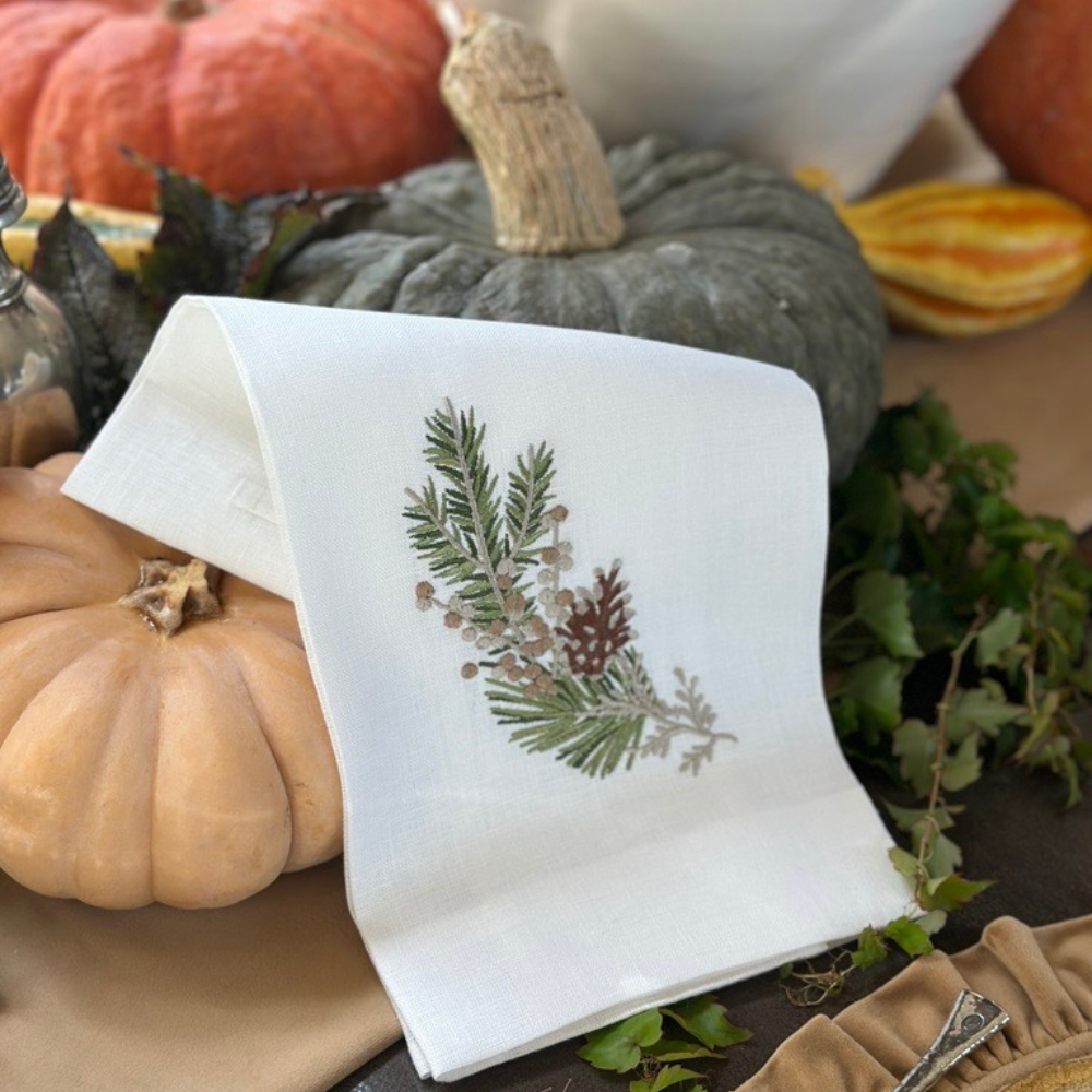 White embroidered kitchen towel with pine branch design on a wooden surface with pumpkins and leaves.
