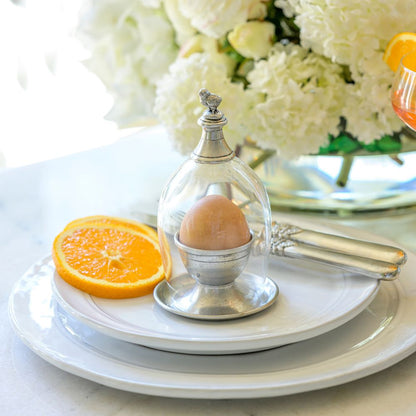 Egg in a pewter holder on a white Firenze plate with an orange slice and flowers in the background