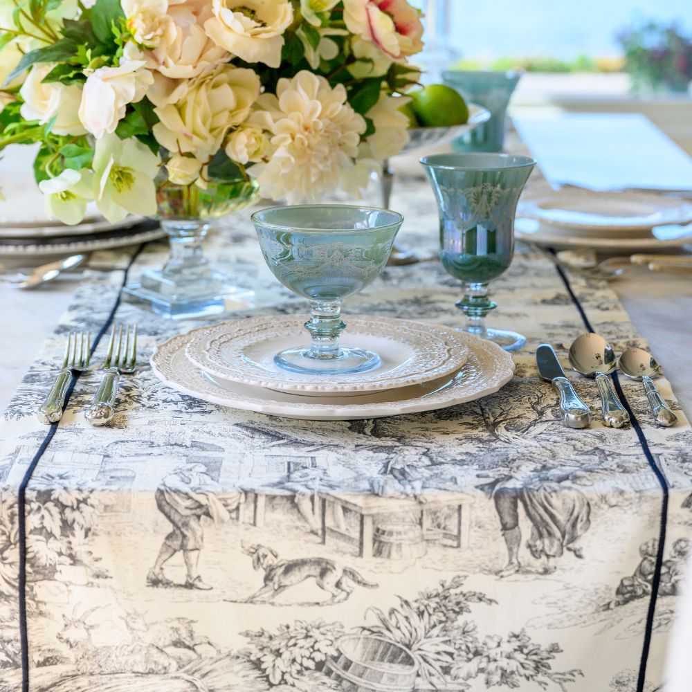 A styled dining table setting featuring a cream and black toile-print table runner, matching plates, blue-green glass goblets, silver flatware, and a large floral centerpiece in the background.