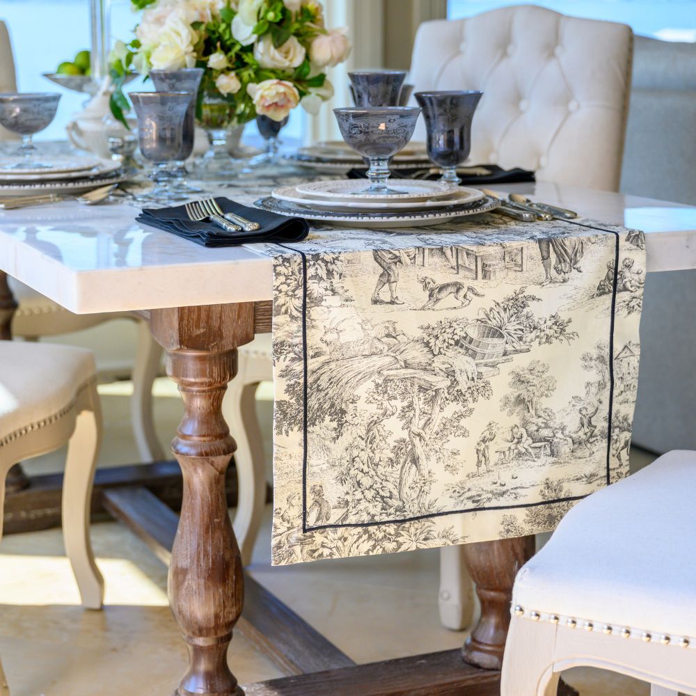 A black and beige toile de Jouy pattern table runner draped over a white marble dining table set with plates, dark glassware, and floral centerpieces.