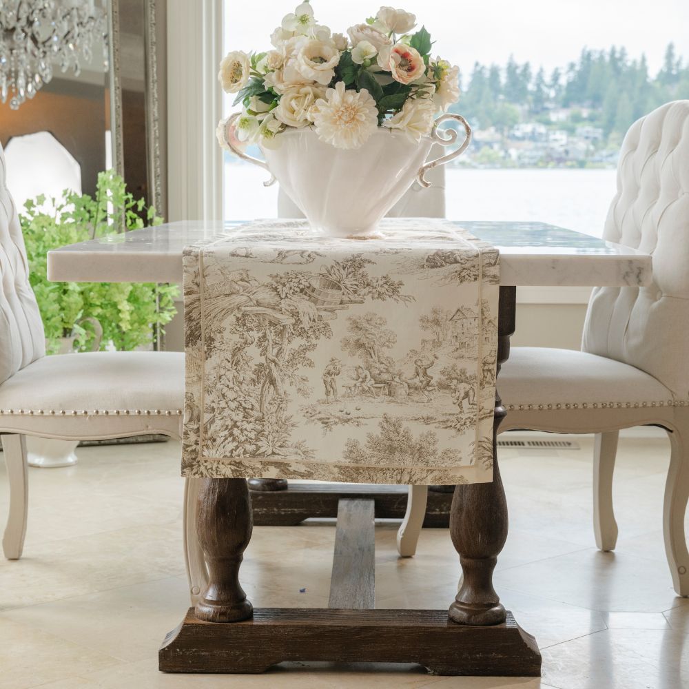 A classic French toile table runner in beige and brown is draped over a dark wood dining table, styled with a large white ceramic pitcher of flowers in the center. The table is surrounded by elegant white tufted chairs in a bright dining room with a window overlooking water.