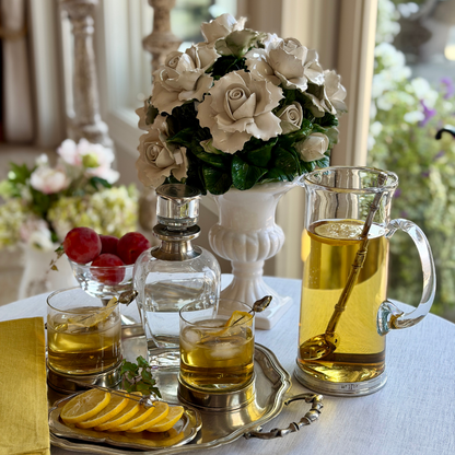 Arte Italica Frankie Martini Pitcher filled with a golden cocktail, styled on a pewter tray with two Portofino Double Old Fashioned glasses, a Scavo decanter, pear cocktail picks, lemon slices, red plums, and a Fresco ceramic white rose topiary