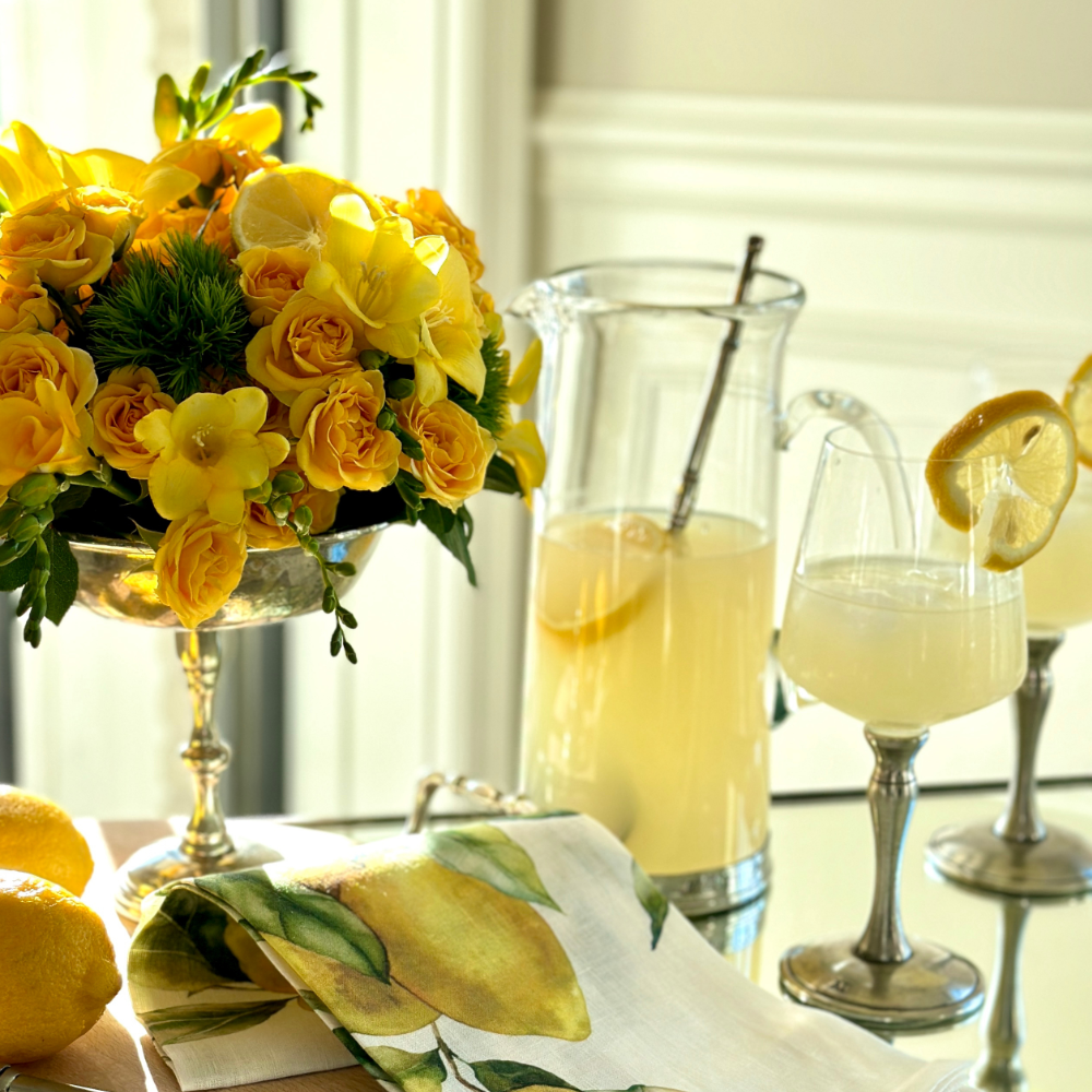 Arte Italica Martini Slotted Mixing Spoon inside a Frankie Martini Pitcher filled with a lemon cocktail, styled with two Verona Martini Glasses with lemon garnishes, fresh lemons, a lemon-print linen napkin, and a yellow rose and orchid arrangement in a pewter compote