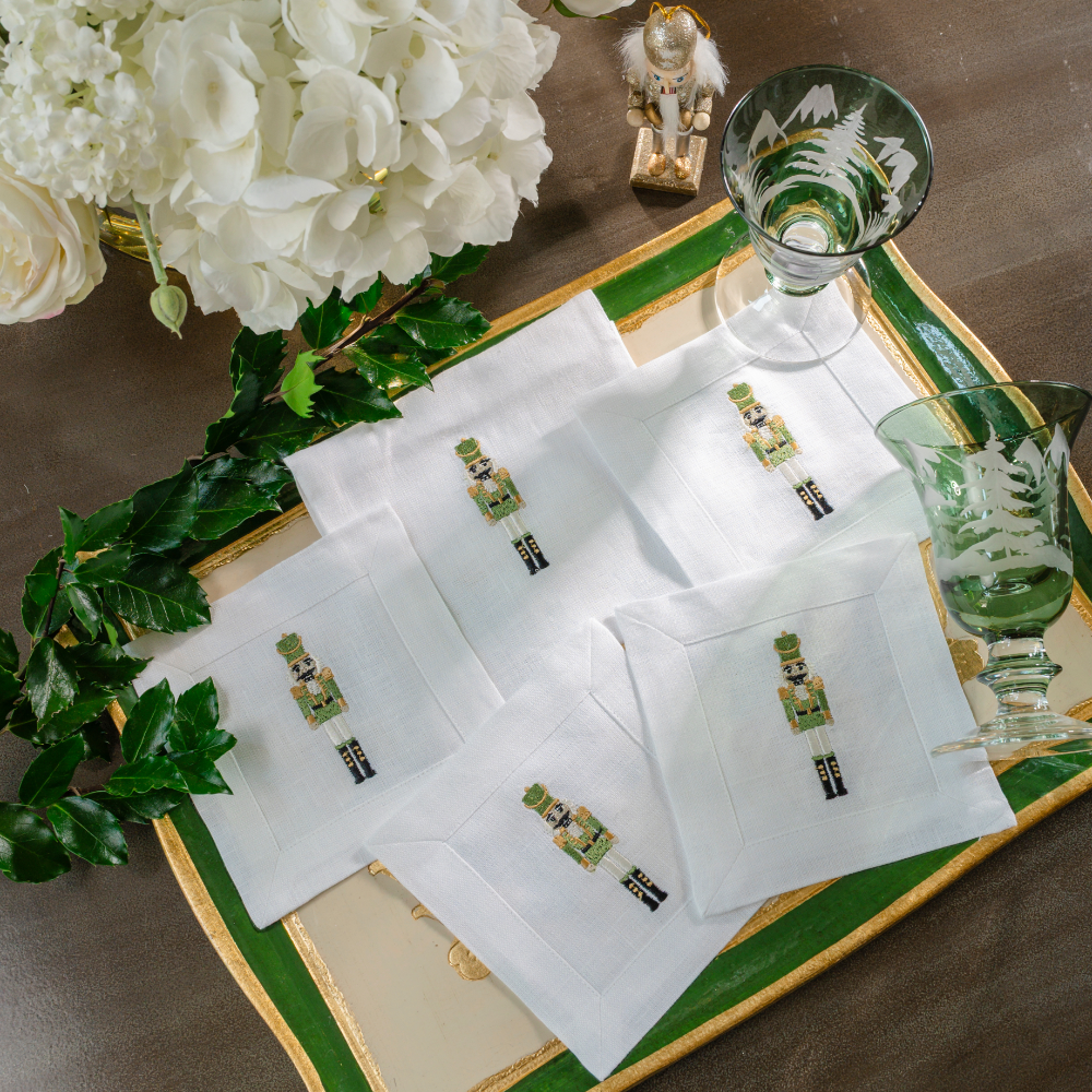 A Florentino green tray with White Cocktail Napkins and pouch embroidered with our green 2026 Nutcracker spread out with greenery and white flowers