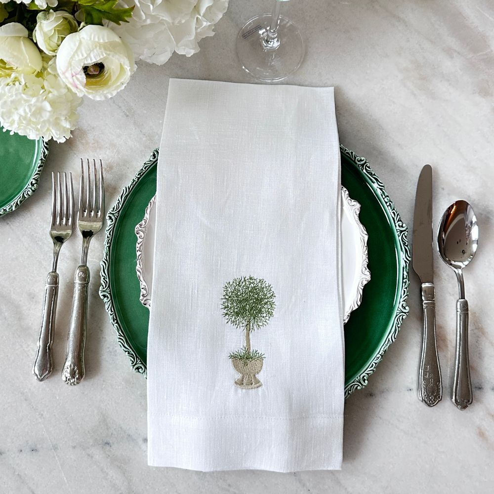 Table setting with green plates, silver cutlery, and a white linen towel with Herb topiary embroidery used as a napkin on a marble surface.