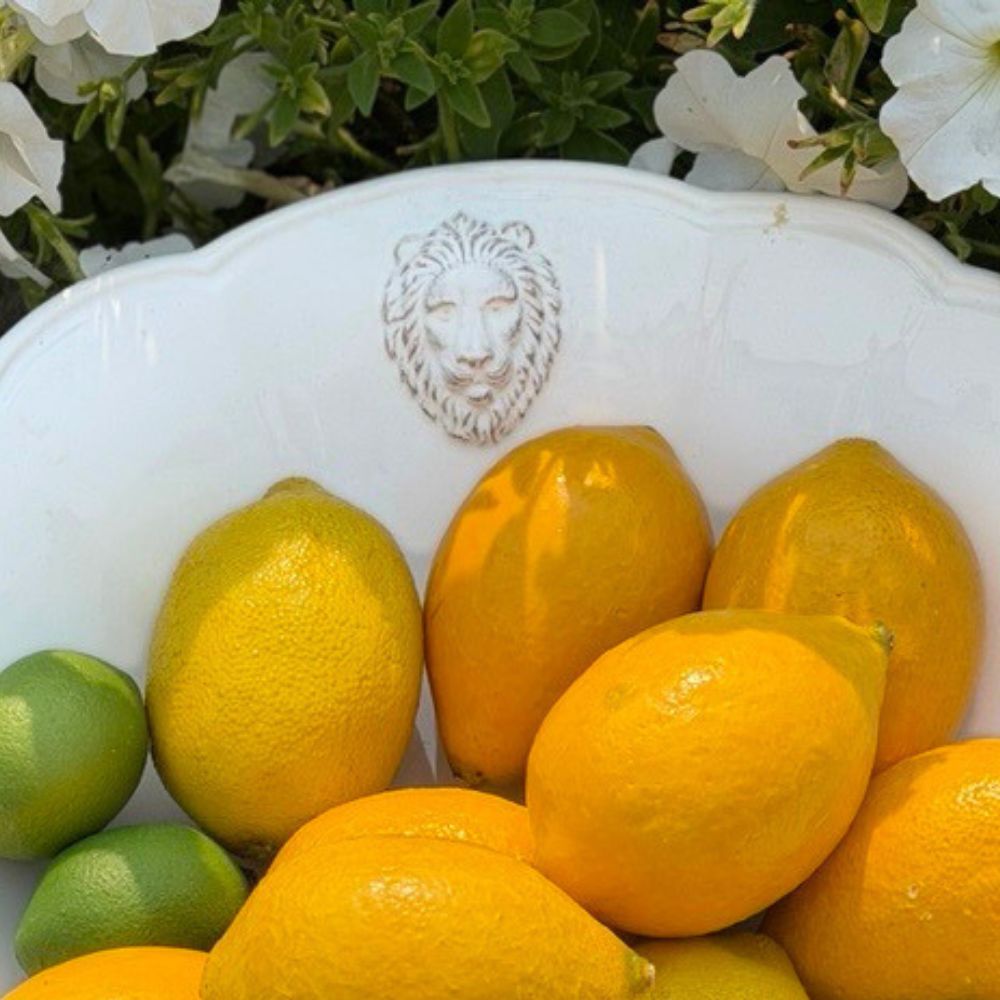 Close up of the raised lion head motif on a Leone Ceramic Serving bowl, filled with bright yellow lemons and a few green limes, set against a background of green foliage and white flowers.