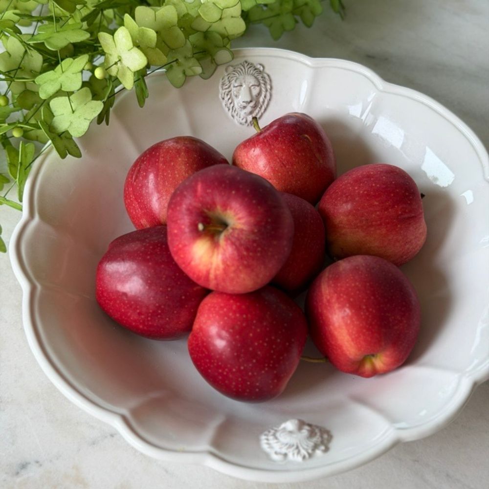 A group of red apples arranged in a white scalloped Leone ceramic serving bowl with decorative lion motifs on the rim, set on a light marble surface with green foliage in the background.