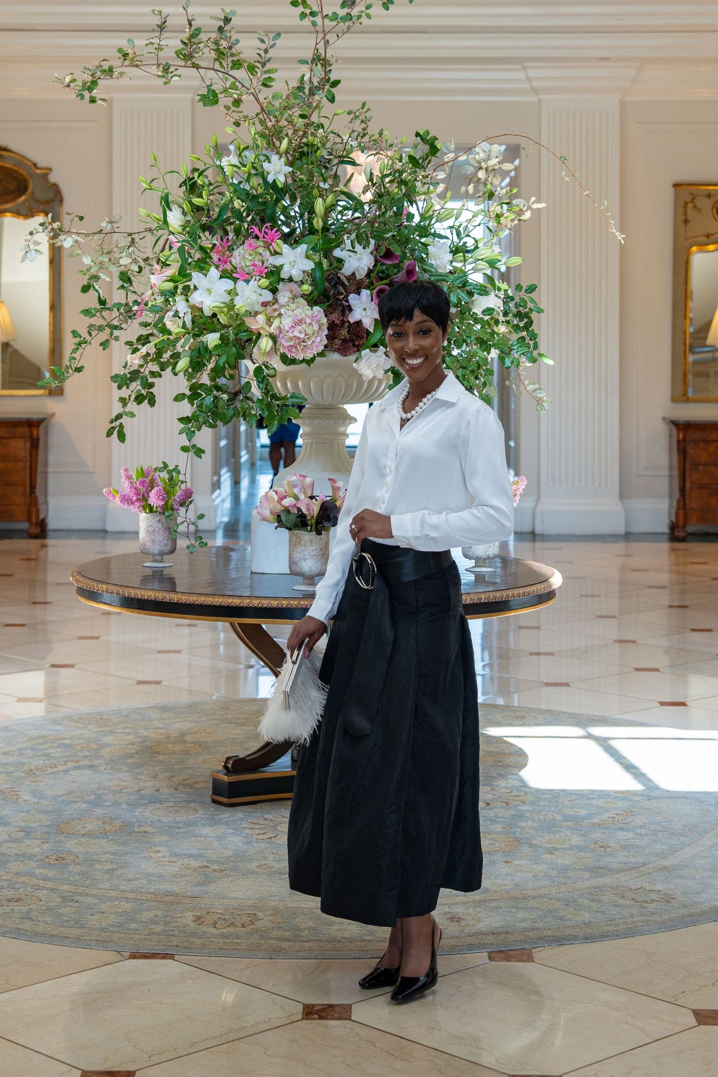 Woman in a white Cameron Viscose blouse and black Lizzy Taffeta skirt standing in a grand hall with floral arrangements.