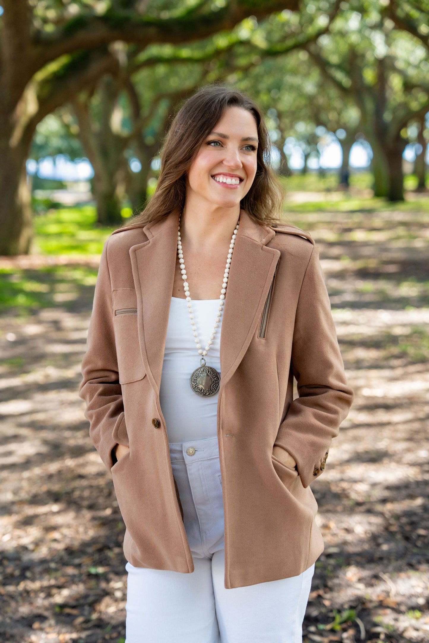 Woman wearing a Camel Colored wool coat and white outfit standing in a park with trees in the background