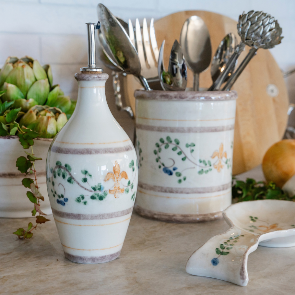 A counter with a Medici ceramic oil bottle, utensil holder and spoon rest with artichokes and a wooden cheese board