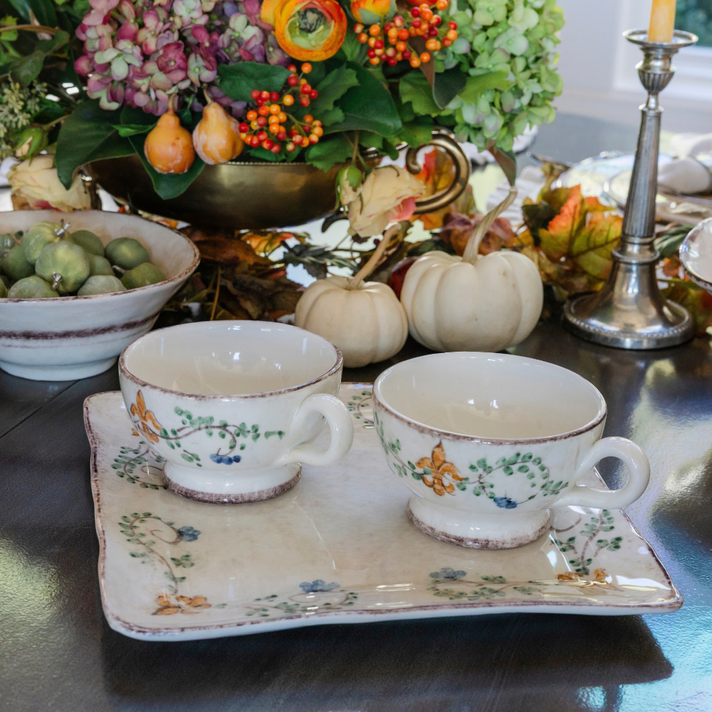 Two Medici grande ceramic mugs on a square charger sitting on a fall table with flowers and small white pumpkins