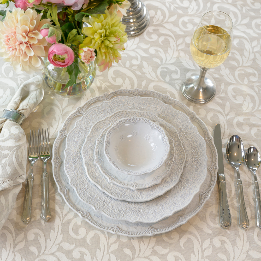 A stack of Merletto Antique white plates on a Crema Toscana Linen tablecloth with flowers and flatware