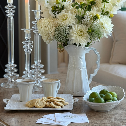 A table with a Merletto scalloped pitcher with white flowers, along side a large oval bowl filled with limes and a square platter holding cookies and mugs with coffee
