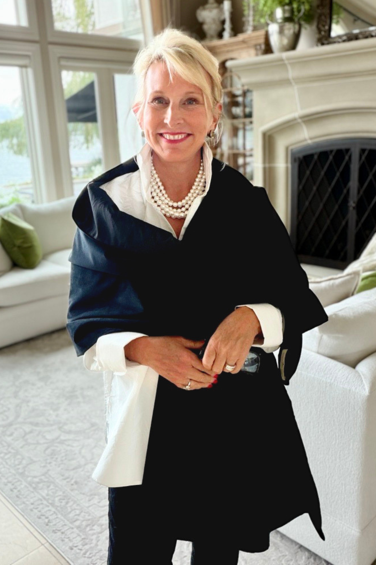 Midnight Wrap in black velvet draped over the shoulders over a white blouse, styled with a layered pearl necklace in a bright, elegant living room with a fireplace