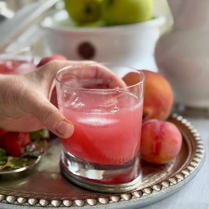Hand holding an Arte Italica Milano Double Old Fashioned glass filled with a pink cocktail over ice, with fresh peaches on a silver tray