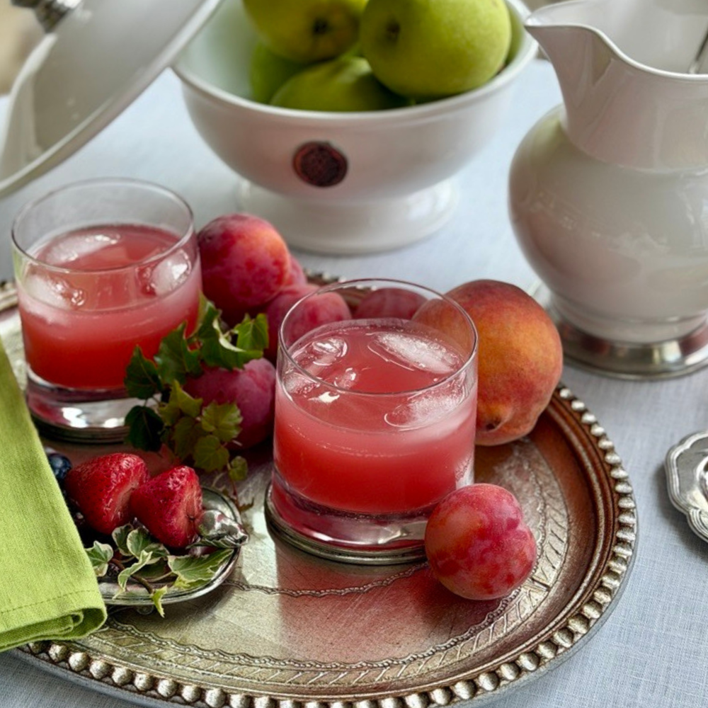 Two Arte Italica Milano Double Old Fashioned glasses filled with a pink fruit cocktail over ice, styled on a silver tray with fresh strawberries, grapes, and peaches