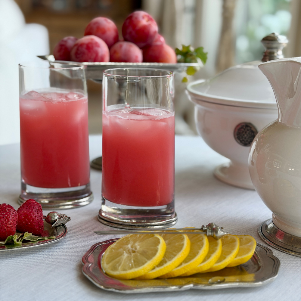 Two Milano double old fashioned glasses with pewter base filled with pink iced lemonade on a white tablecloth, with lemon slices, strawberries, plums, and a Tuscan Arte Italica pieces.