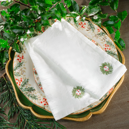 Two folded white linen napkins embroidered with a mini wreath with red berries on a Natale plate with holly