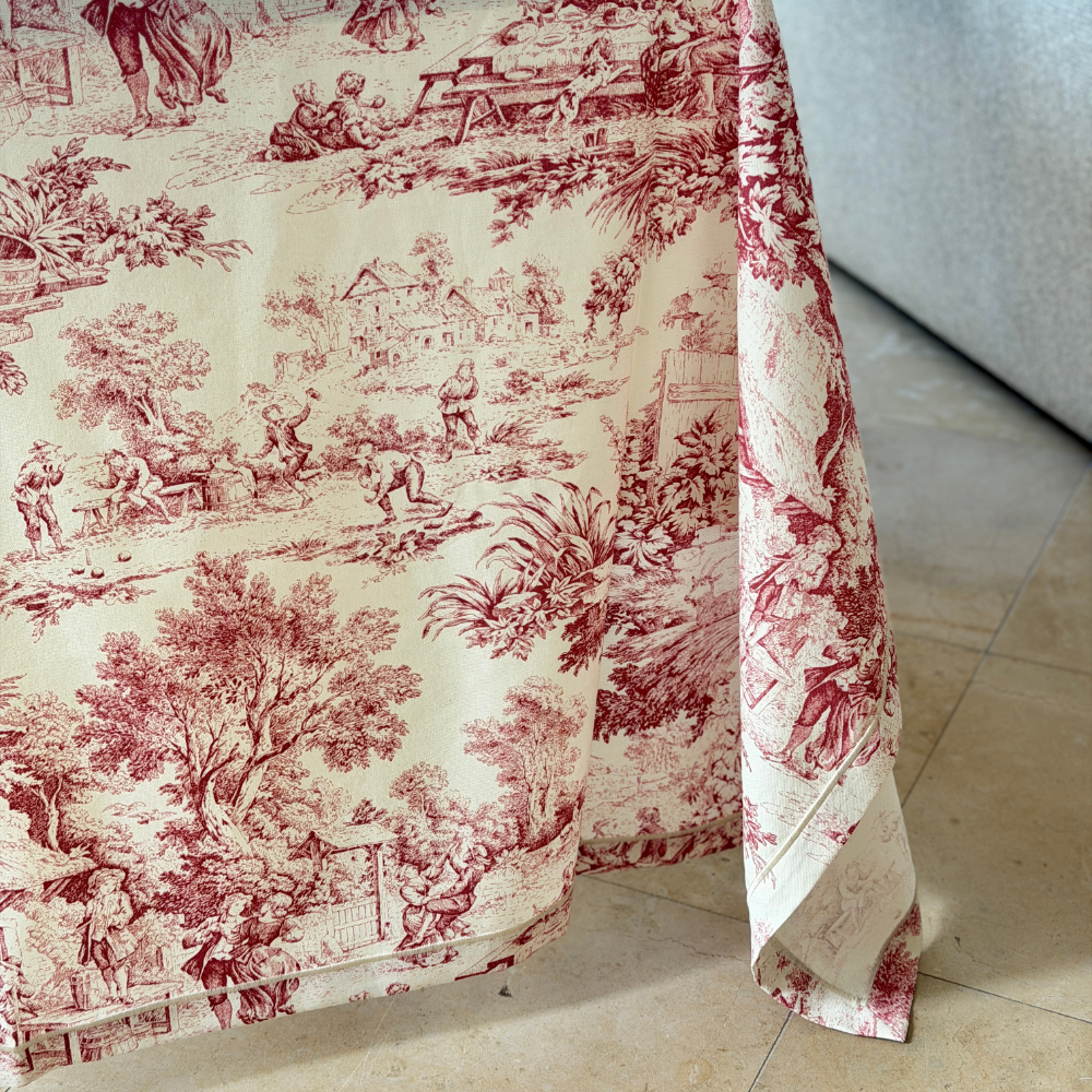 Close-up detail of the Arte Italica Montefalco Toile Cotton Tablecloth in red, showing classic toile pastoral scenes with figures, trees, and countryside cottages on an ivory cotton ground