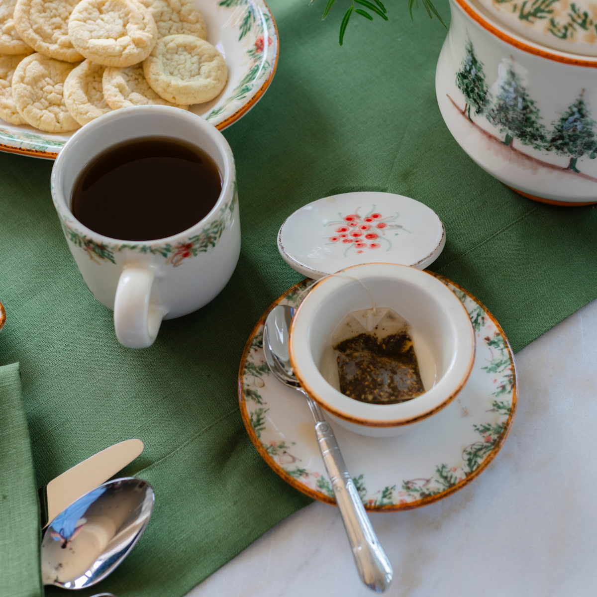 A top view of the Natale Italian ceramic tea infuser mug sitting next to the infuser with a teabag in it on a bread plate with a spoon