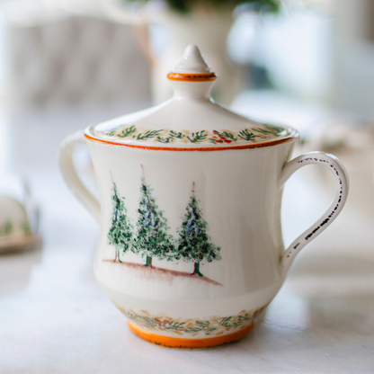 A two handled cookie jar with lid in the Arte Italica Natale pattern with 3 trees painted on it close up on a blurred background