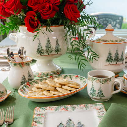 A table set with Natale ceramic pieces on a evergreen linen tablecloth with an oval dish with sugar cookies and cachepot with red roses 